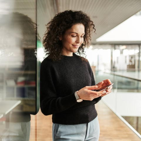 Frau lehnt an Glasfront und blickt lächelnd auf ihr Smartphone