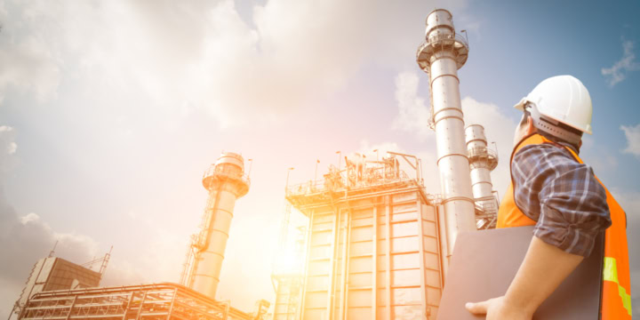 Mann in Schutzkleidung mit Laptop unter dem Arm blickt auf eine industrielle Anlage