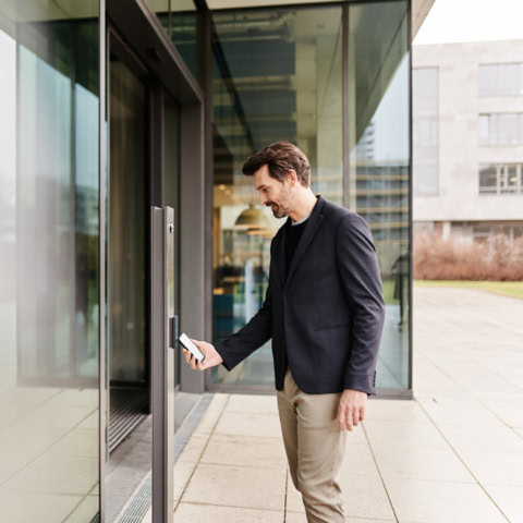 Mann vor Gebäudeeingang hält Smartphone mit Key App an Interflex IF-800 Zutrittsterminal