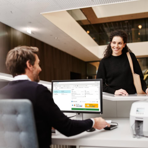 Mann beschreibt IF-73 mit Zutrittsrechten mit Interflex-Besuchermanagement-Software während Frau lächelnd am Empfang steht