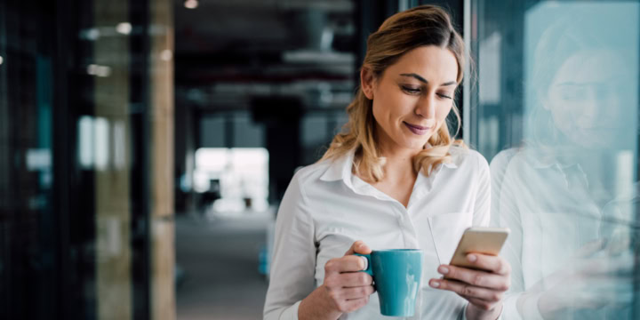 Frau lehnt an einer Glasfront, in den Händen eine Tasse und ein Smartphone, auf das sie lächelnd blickt