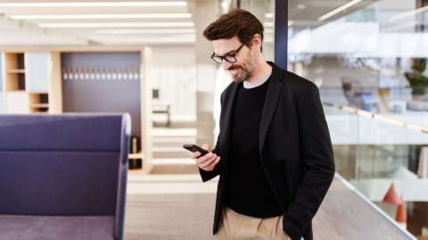 Mann im modernen Firmengebäude blickt lächelnd auf sein Smartphone