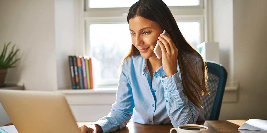 Woman at desk with smartphone to her ear smiles at her laptop.